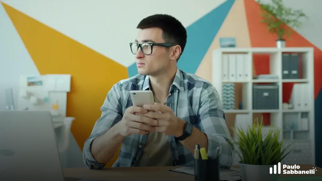 Homem de óculos segura celular sentado à mesa com notebook, parede colorida geométrica ao fundo