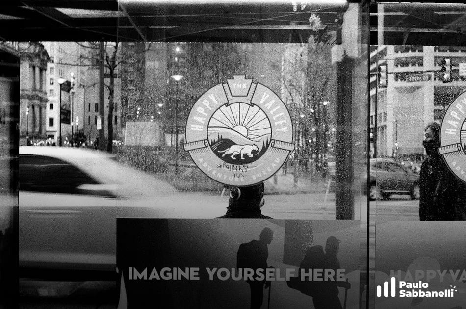 Vitrine com o texto Imagine Yourself Here refletindo rua movimentada, foto em preto e branco