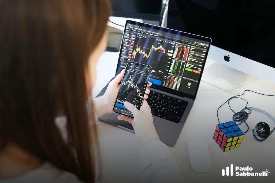 Mulher observa celular e notebook com gráficos financeiros, cubo mágico e fone ao lado
