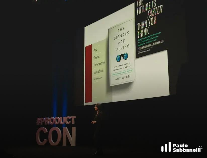 Palestrante em evento ProductCon com capas de livros projetadas em telão atrás
