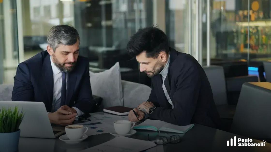 Dois consultores de terno conversam à mesa com café, cidade ao fundo pela janela