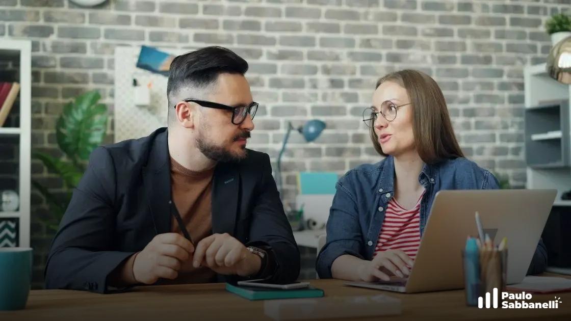 Homem e mulher conversam sentados à mesa com notebook em escritório de tijolo aparente