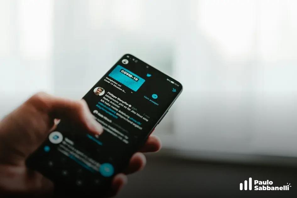 Mão segurando um celular com o aplicativo do Twitter em modo escuro exibindo um tuíte sobre covid