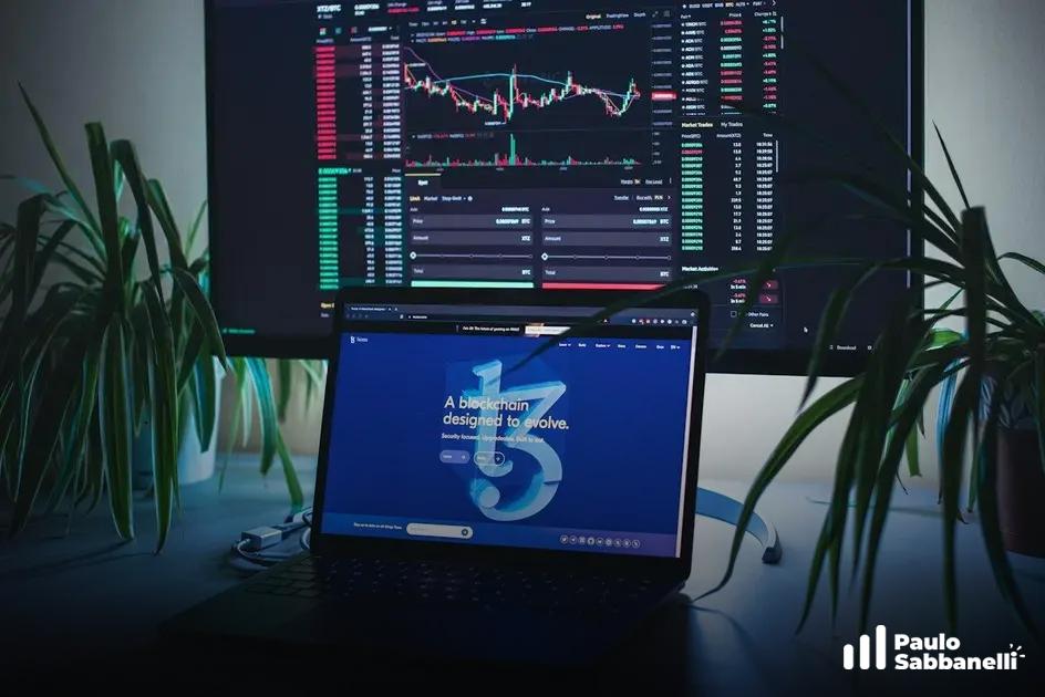 Mesa com monitor de gráficos financeiros e notebook aberto em site sobre blockchain, cercados de plantas