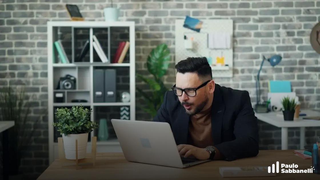 Homem de blazer digita concentrado no notebook, com expressão de foco, em escritório com parede de tijolinhos