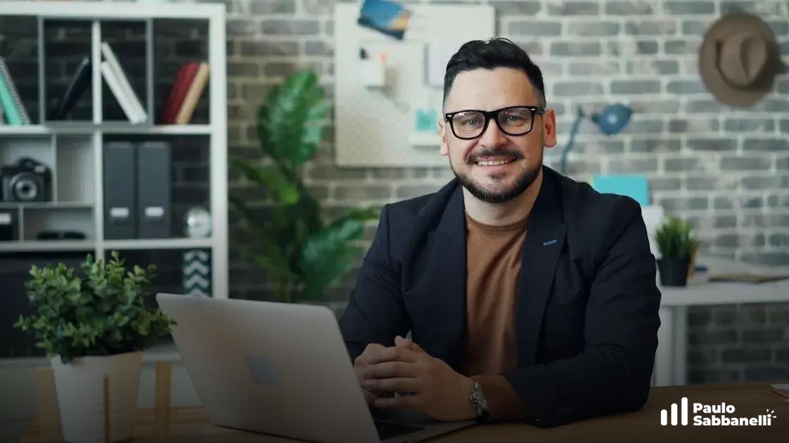 Homem de blazer escuro sorri sentado à mesa com notebook, em escritório com parede de tijolinhos e plantas