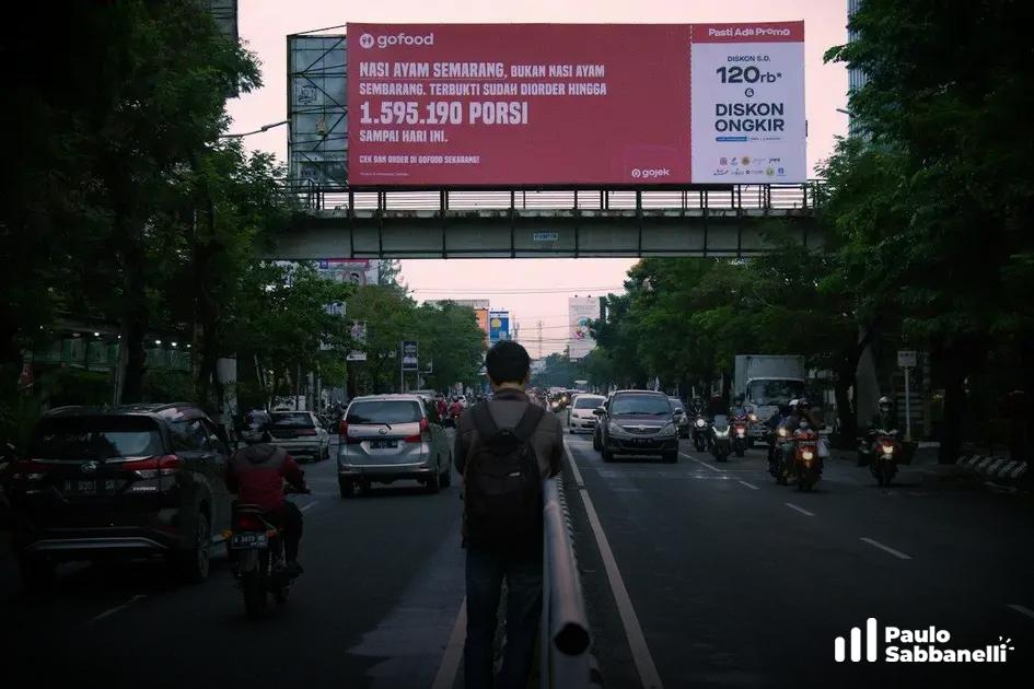 Homem caminha por avenida movimentada com painel publicitário de comida ao fundo