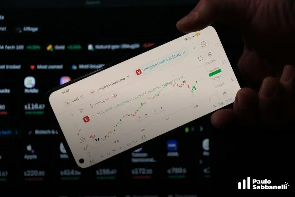 Mão segura celular exibindo gráfico de investimentos em alta, com monitor de cotações ao fundo