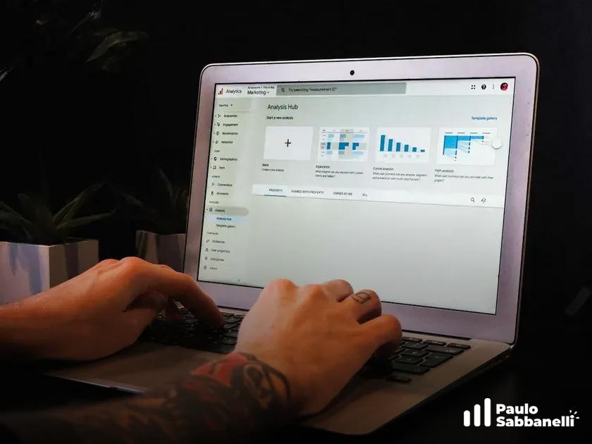 Mãos tatuadas digitam em notebook com painel de análise e gráficos de barras na tela