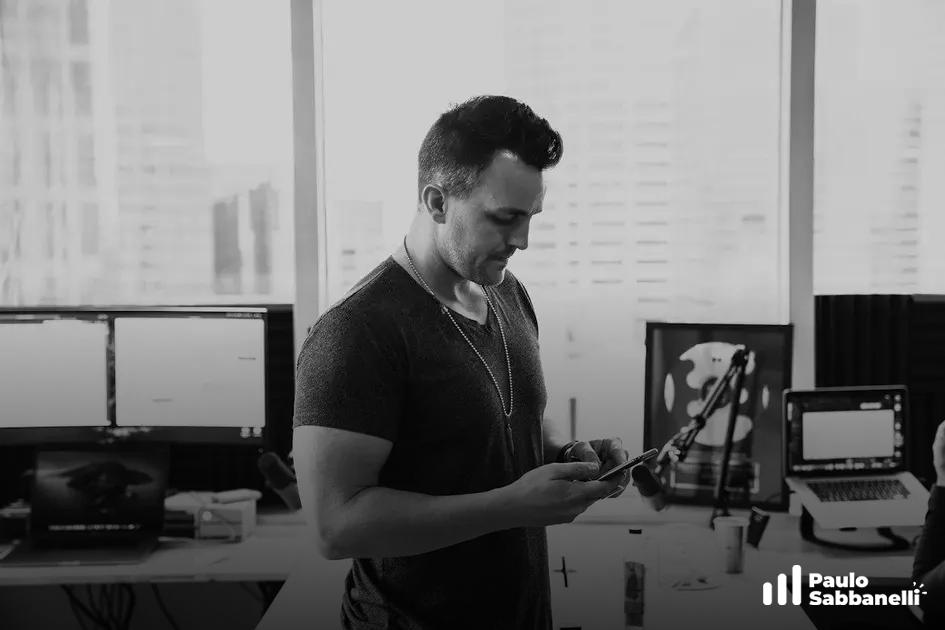 Foto em preto e branco de homem mexendo no celular em estúdio com monitores e equipamentos de gravação