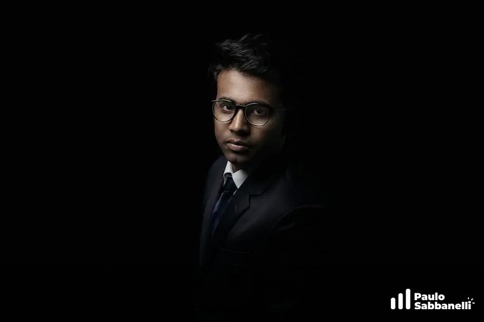 Retrato em tom escuro de um jovem de terno e óculos, iluminado apenas no rosto