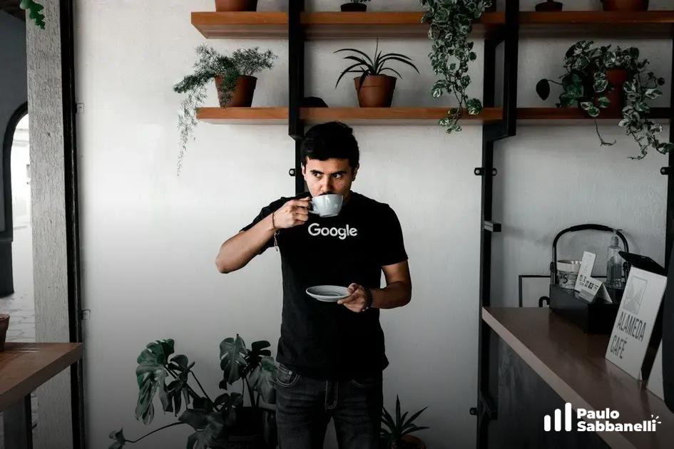 Homem bebendo café em uma xícara com a marca Google, em cafeteria com plantas ao fundo